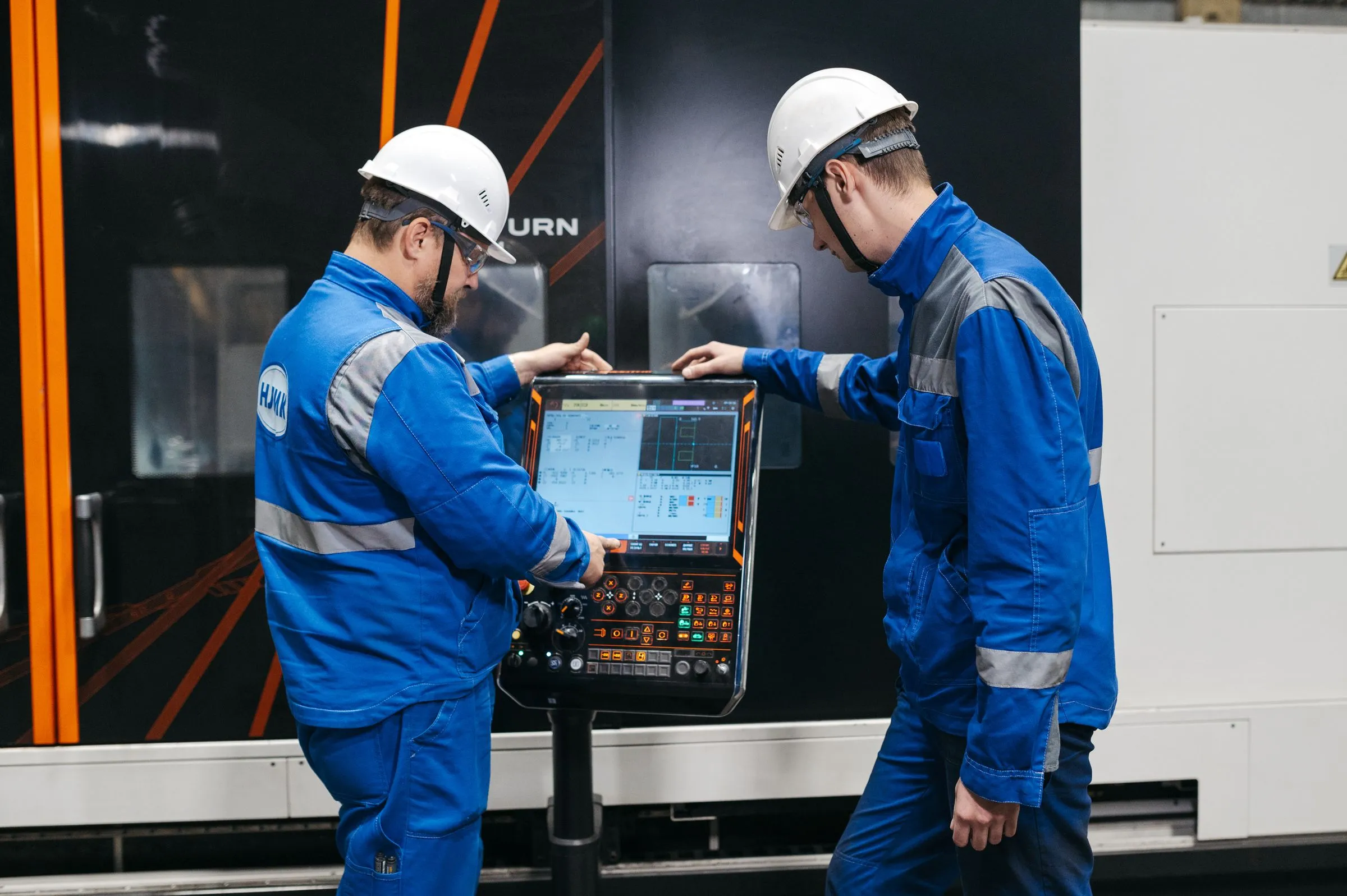 Two technicians in hard hats working at a CNC machine’s touchscreen control panel