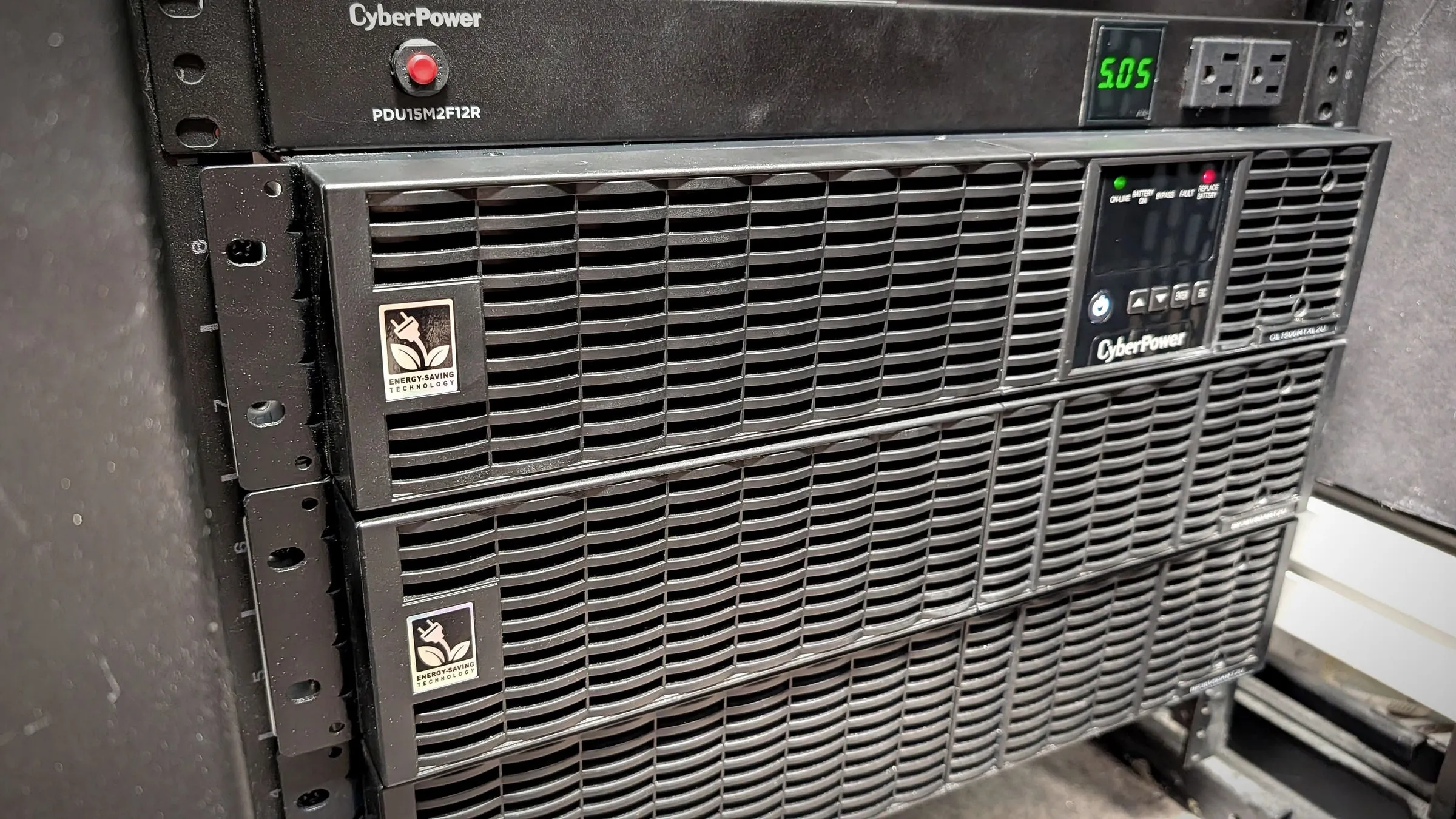 Rack-mounted UPS and battery pack below a power distribution unit, with the red replace-battery light lit on the UPS panel